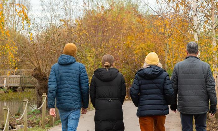 A group walk through an autumnal scene alongside a pond. Yellow and orange leaves against the grey autumn sky.
